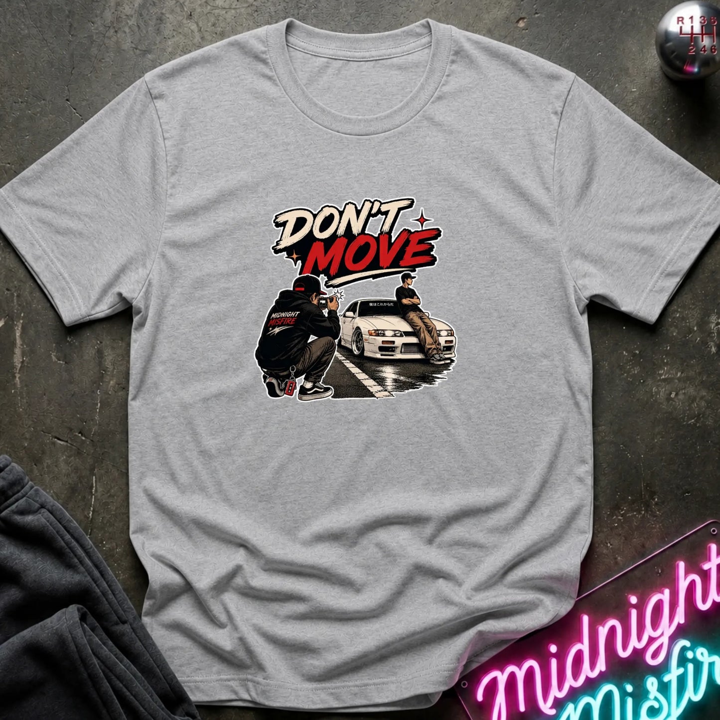A light gray crewneck t-shirt with a graphic print on the front. The graphic features the text 'DON'T MOVE' in stylized white and red lettering above an illustration of a person crouching next to a white sports car on a wet street.