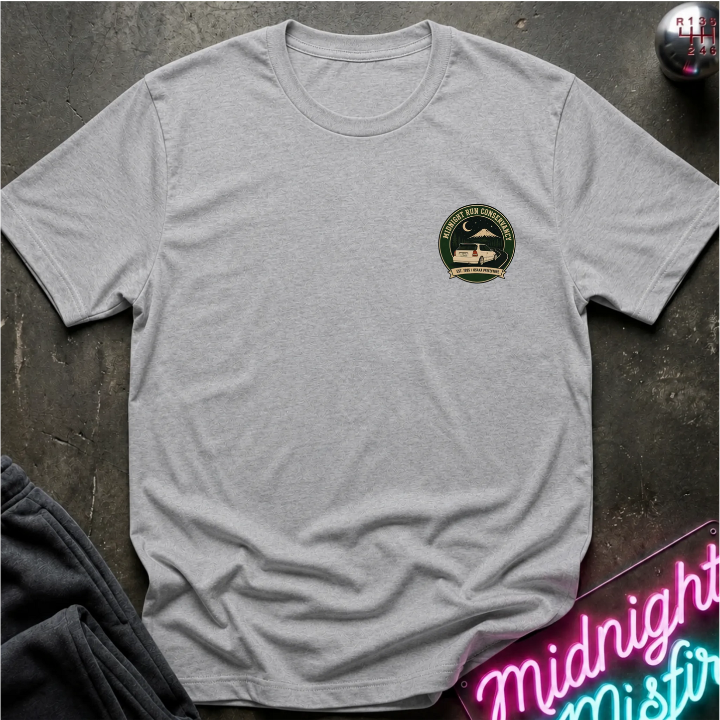A light gray heather short-sleeve crewneck t-shirt with a circular green and black chest graphic, styled flat-lay on a dark textured surface with a neon sign and dark pants nearby.