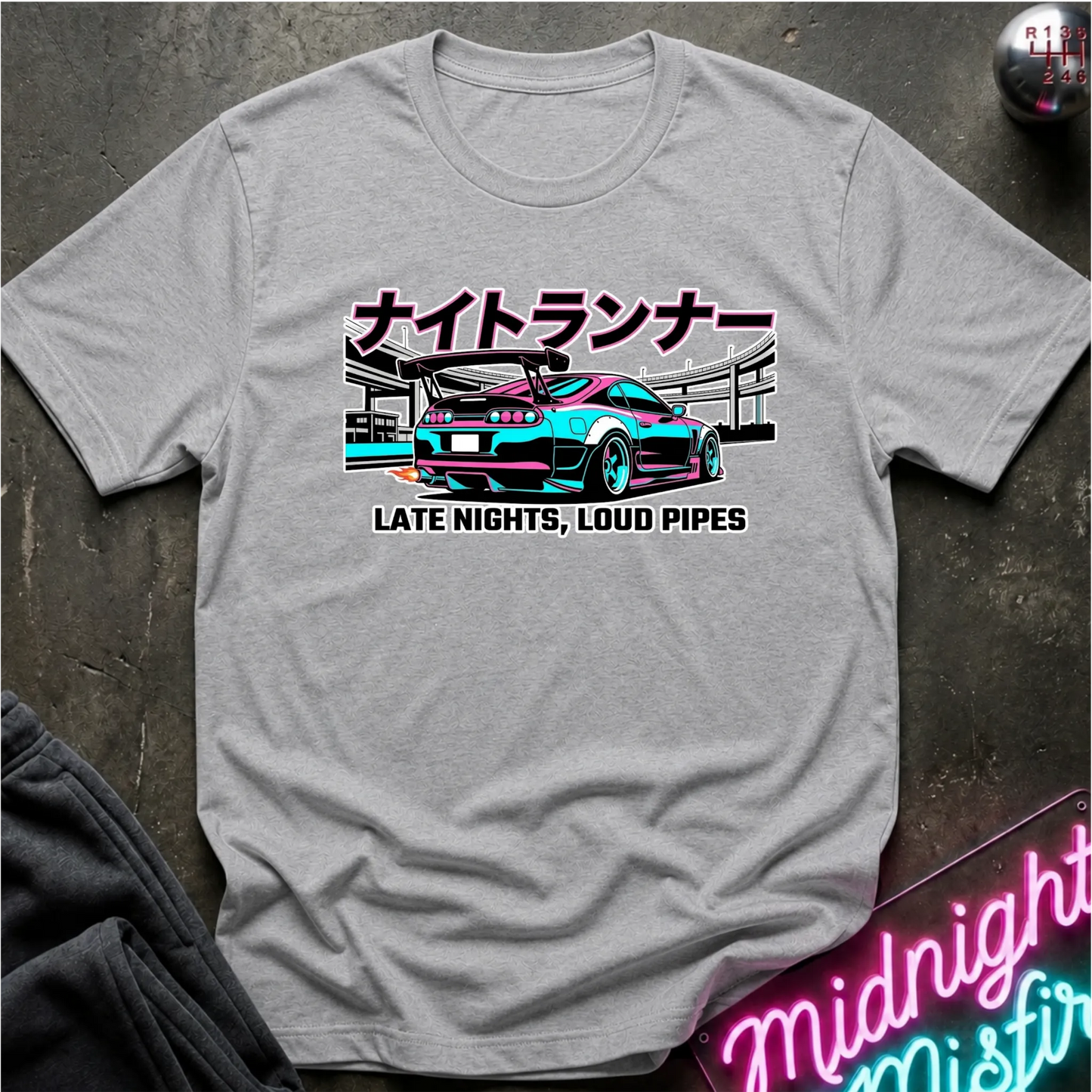 A light gray heather crewneck t-shirt featuring a graphic of a teal and pink sports car with Japanese text above and 'LATE NIGHTS, LOUD PIPES' below. The shirt is laid flat on a dark textured surface with a neon sign and dark pants partially visible.