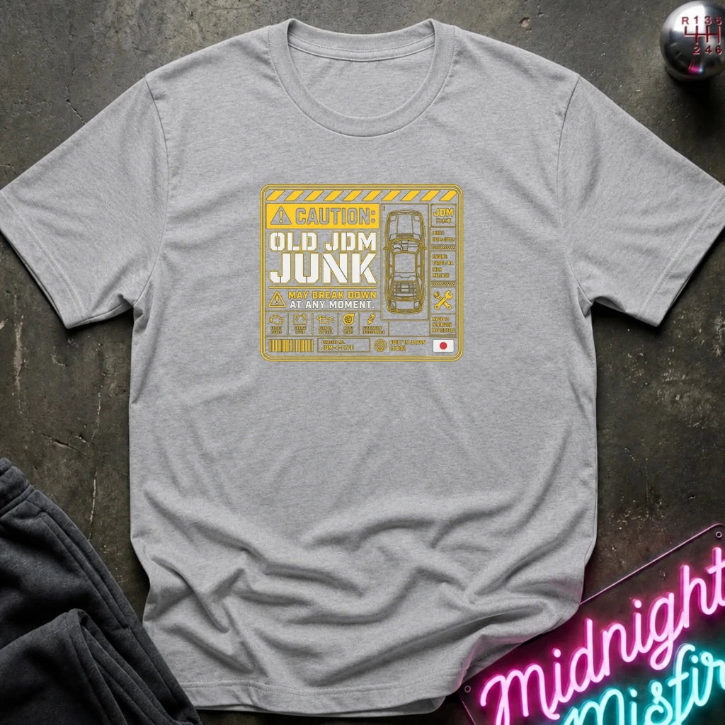 A heather gray short-sleeve t-shirt with a vintage-style yellow and white graphic on the chest reading 'Old JDM Junk' and 'The Awakening.' The shirt is laid flat on a dark textured surface with a neon sign and dark pants partially visible as styled props.