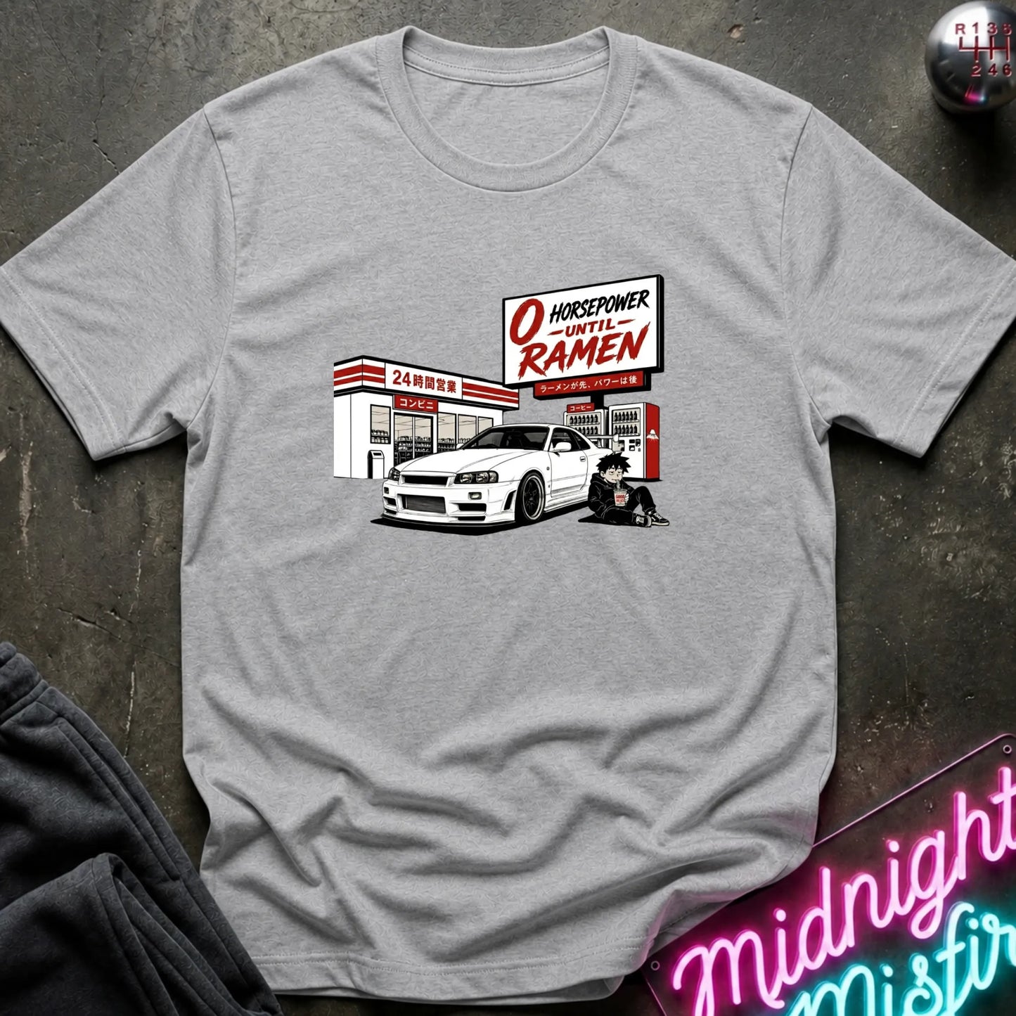A heather gray short-sleeve t-shirt with a graphic print of a white sports car in front of a store , styled flat on a dark textured background with a neon sign and dark fabric in the corners.
