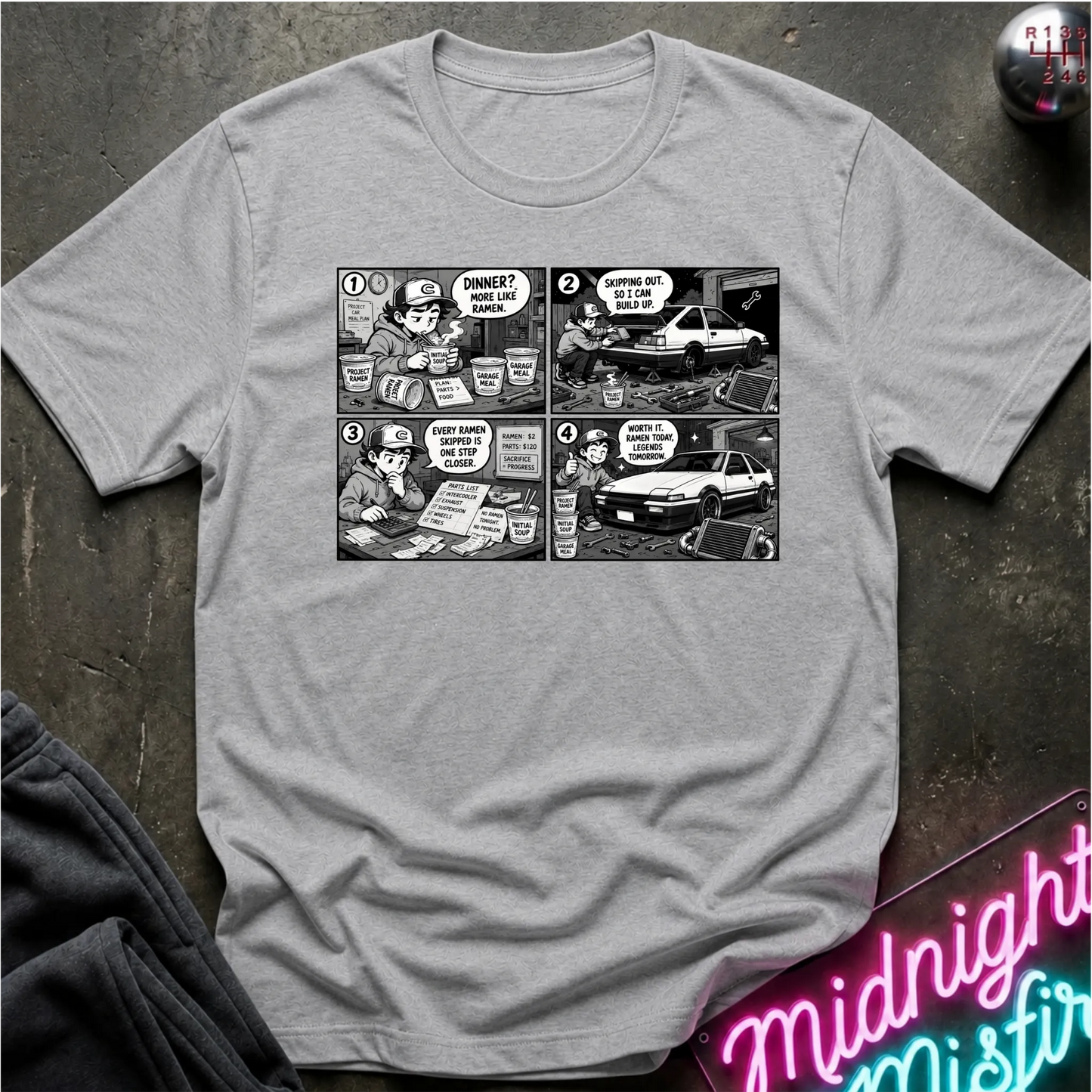 A light gray heather short-sleeve crewneck t-shirt featuring a black and white comic strip graphic on the chest, styled in a flat-lay on a dark textured surface with a neon sign and dark pants partially visible.