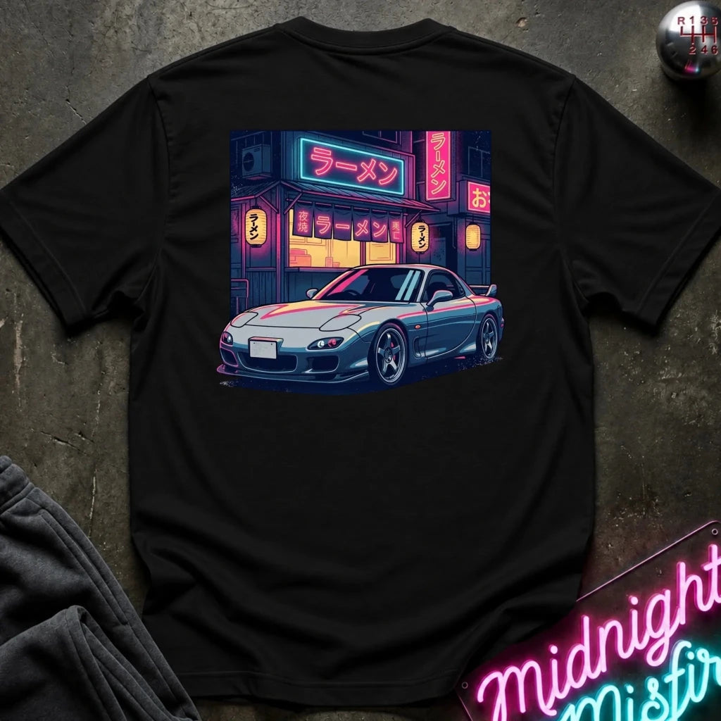 A black short-sleeve t-shirt laid flat on a dark textured surface, featuring a colorful graphic print of a blue sports car parked in front of a neon-lit ramen shop.