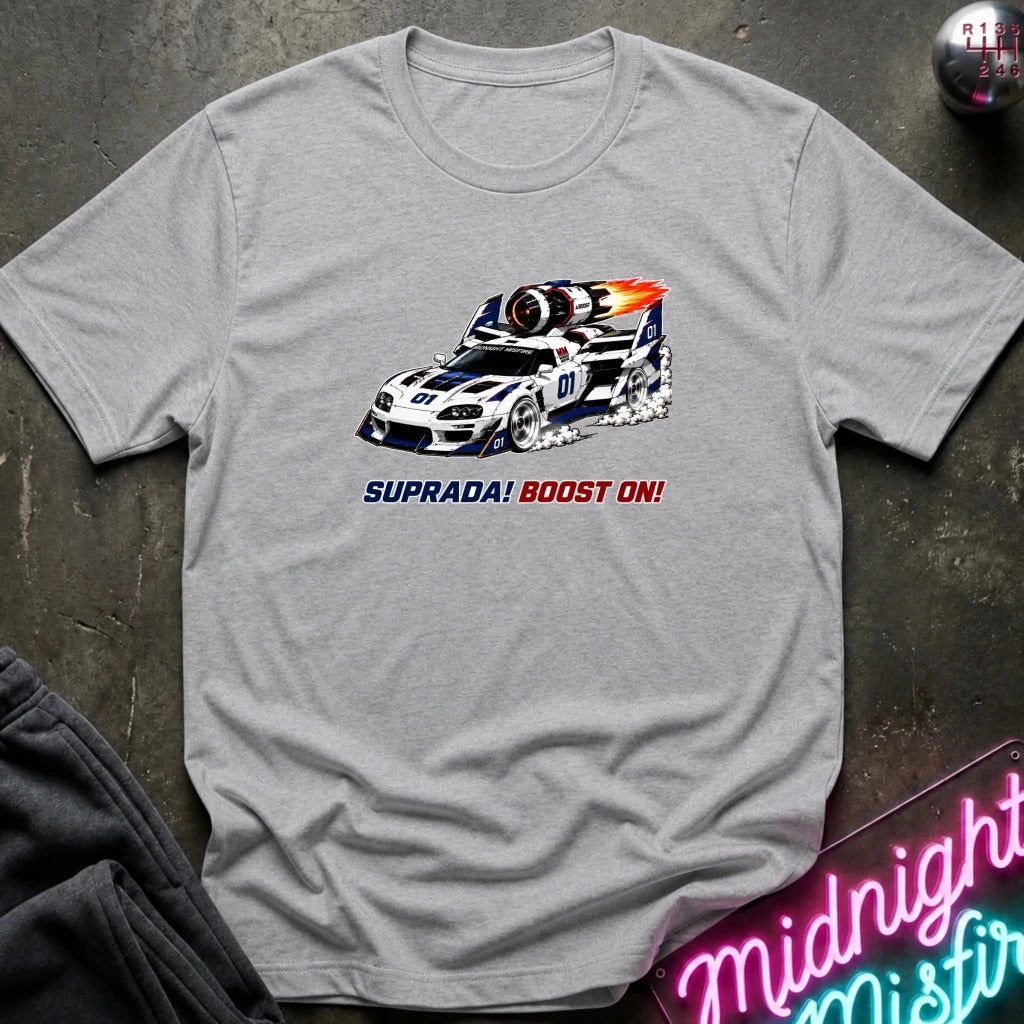 A light gray crewneck t-shirt featuring a graphic of a white and blue race car with a red and orange flame, and the text 'SUPRADA! BOOST ON!' printed below it. The shirt is displayed flat on a dark textured surface with a neon sign and dark pants partially visible.