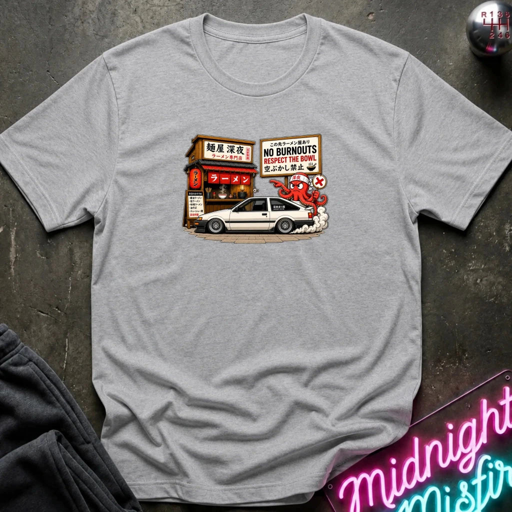 A heather gray short-sleeve t-shirt laid flat on a dark textured surface, featuring a graphic print of a white sports car parked in front of a Japanese street shop with signs reading 'NO BURNOUTS'.