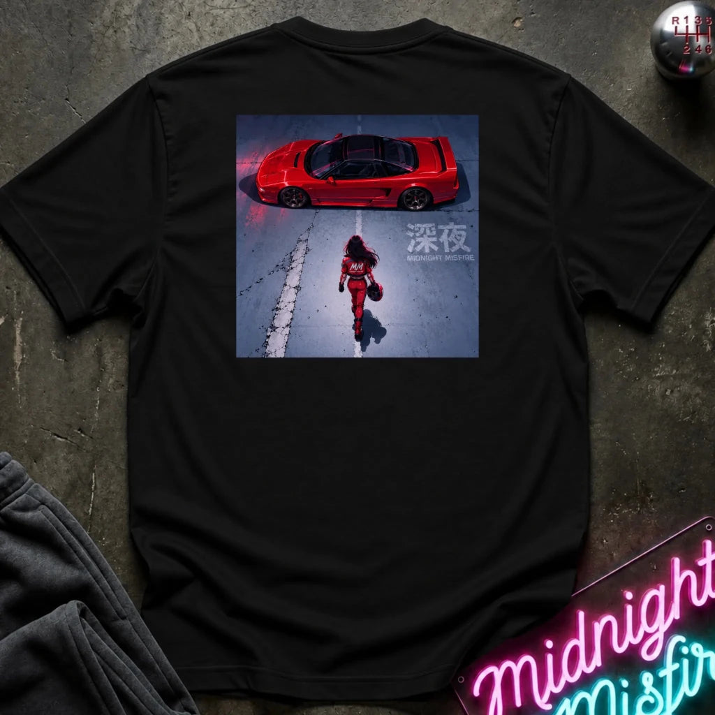 black short-sleeve t-shirt laid flat on a dark textured surface, featuring a large rectangular graphic print on the back. The graphic shows a red sports car in a snowy scene with a person walking