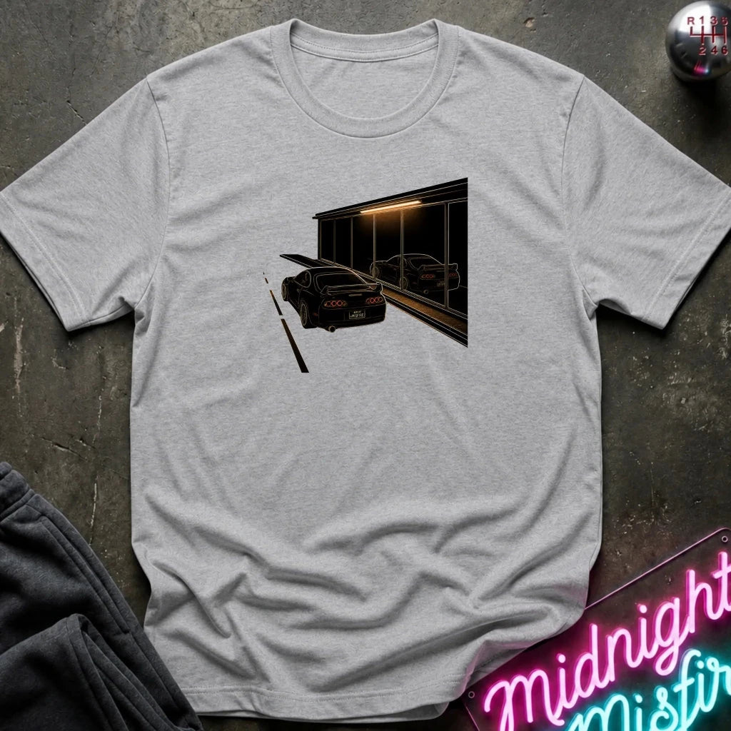 A light heather gray short-sleeve t-shirt laid flat on a dark textured surface, featuring a graphic print of a black sports car in front of a dark building with warm light. 