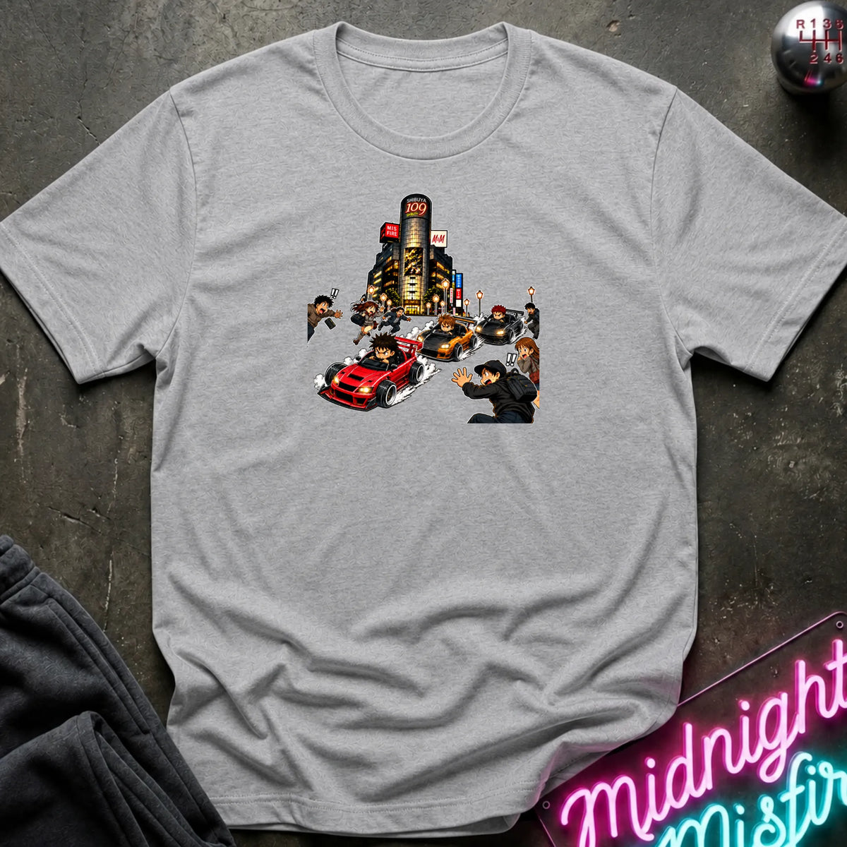 A heather gray short-sleeve crewneck t-shirt with a graphic print of a city street scene featuring a red sports car and small figures, laid flat on a dark textured surface with a neon sign and dark pants partially visible.