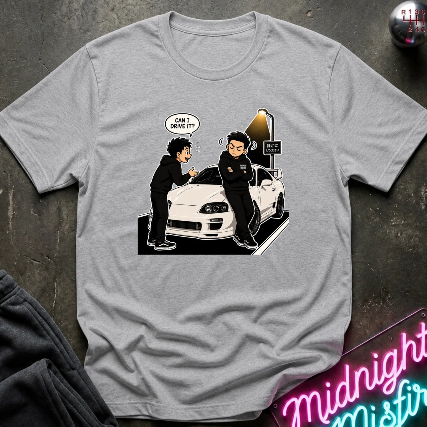 A light gray heather crewneck t-shirt featuring a cartoon graphic of two men in black standing next to a white sports car, with one speech bubble reading 'CAN I DRIVE IT?'. The shirt is displayed flat on a dark textured surface with a neon sign and dark sweatpants partially visible.