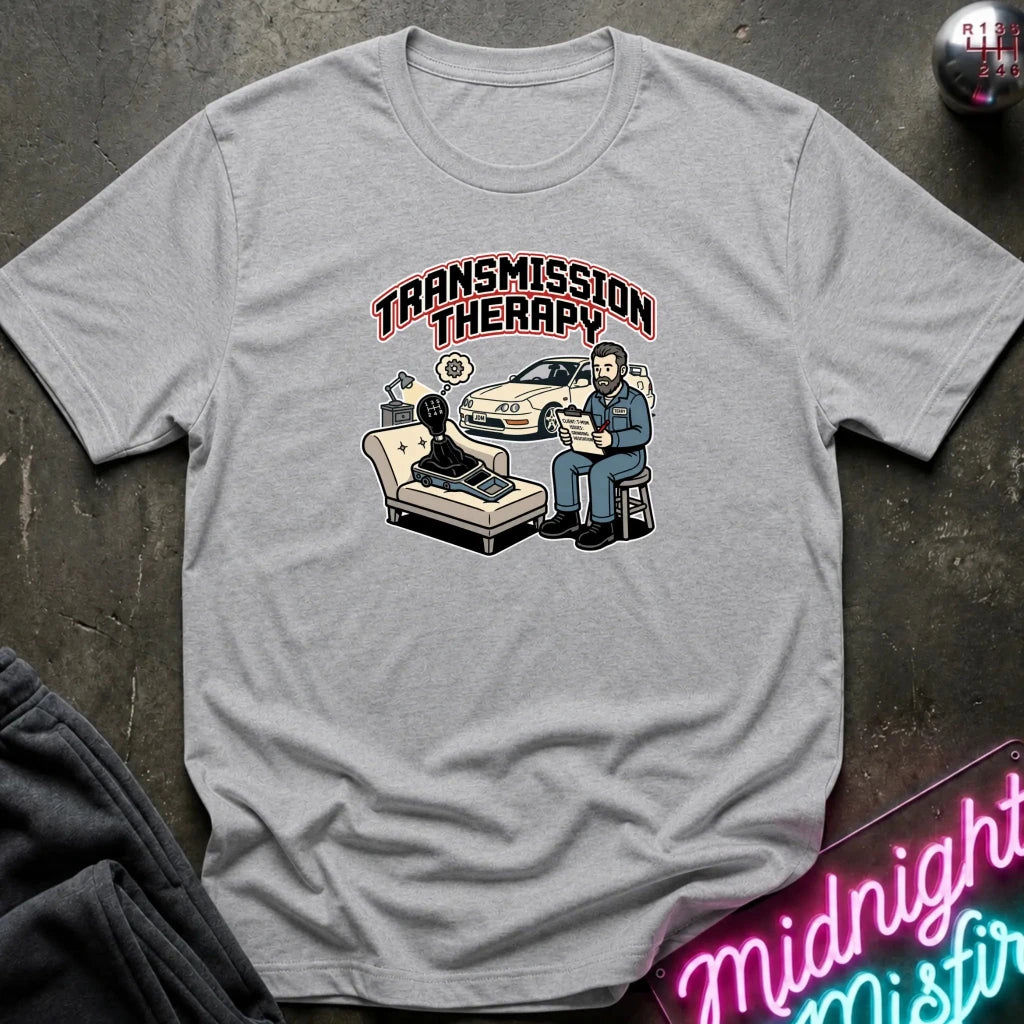 A light gray crewneck t-shirt with a 'Transmission Therapy' graphic print, laid flat on a dark textured surface with a neon sign and sweatpants partially visible.