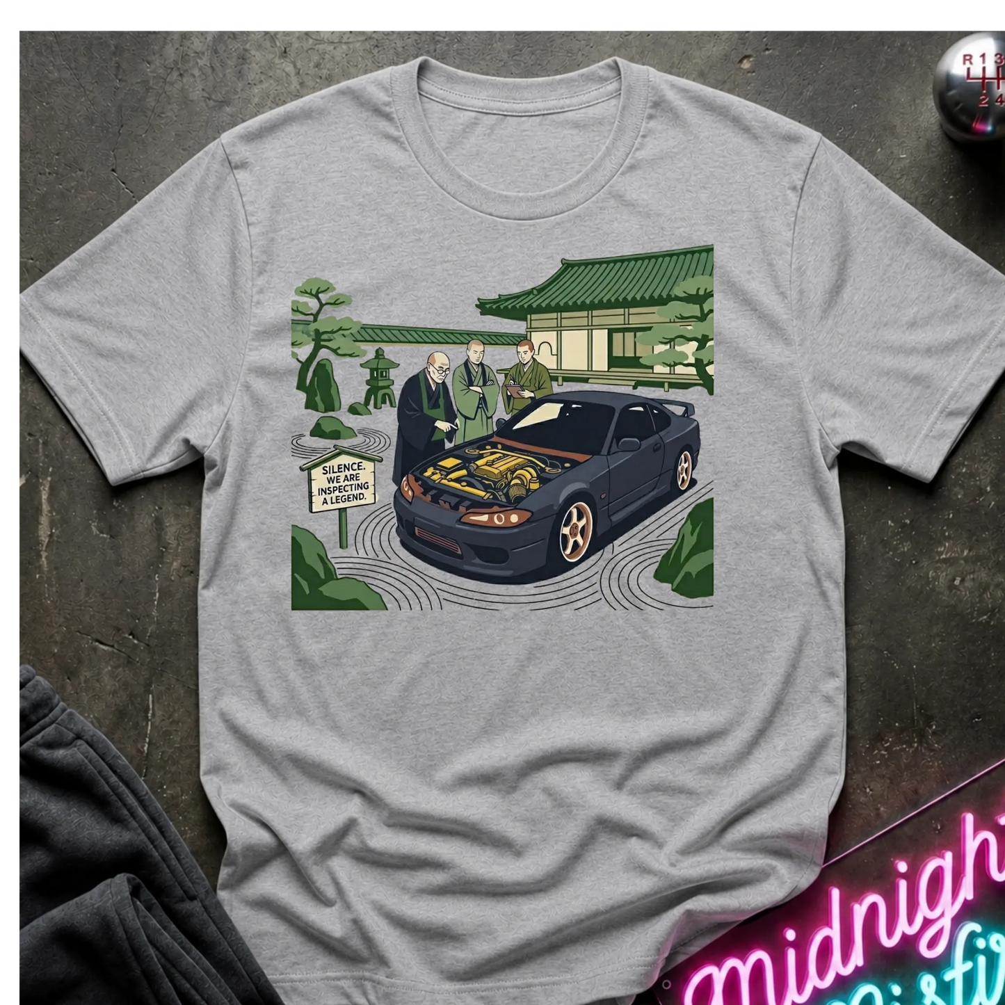 Gray  short-sleeve graphic t-shirt laid flat on a dark textured surface. The graphic shows a black sports car with its hood open, surrounded by two men in suits, a Japanese-style building, and green trees.