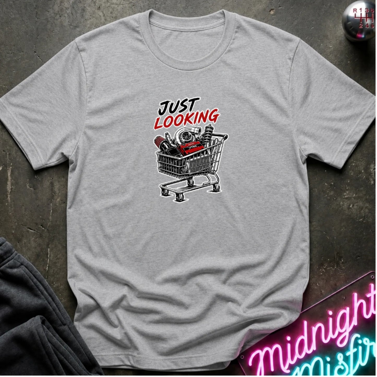 A light gray heather crewneck t-shirt laid flat on a dark textured surface. The shirt features a graphic print of a shopping cart filled with various items and the text 'JUST LOOKING' in black and red.