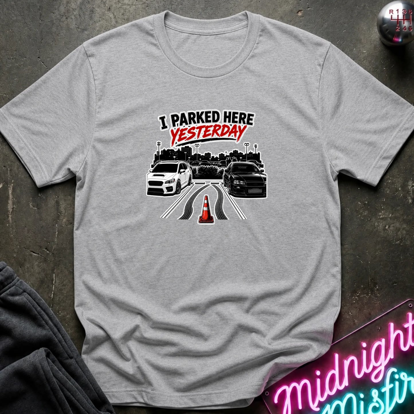 A heather gray short-sleeved t-shirt  that has 2 cars parked in a space in between empty space with skid marks.