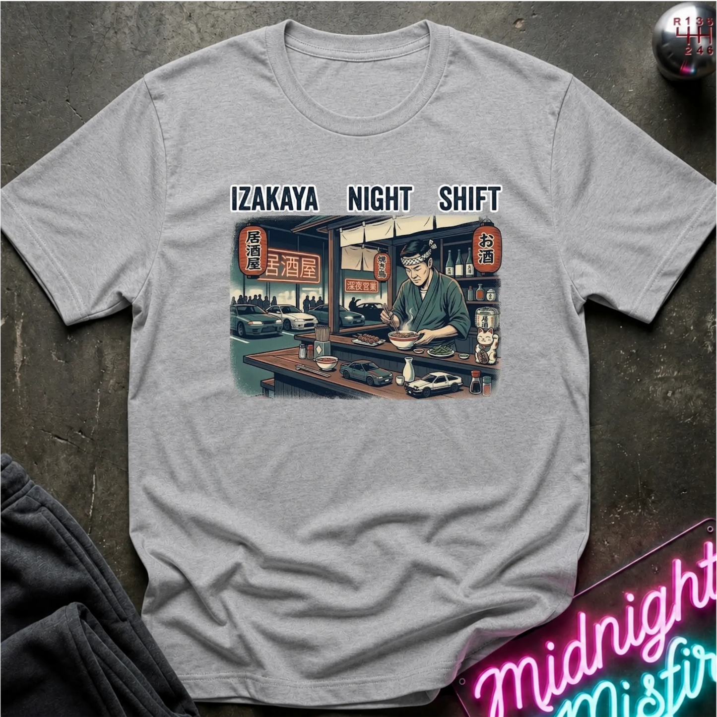 A light gray crewneck t-shirt featuring a graphic print of an employee working a night shift at an izakaya restaurant, with the text 'IZAKAYA NIGHT SHIFT' above the illustration. The t-shirt is displayed flat on a dark textured surface with a neon sign and dark fabric in the background.