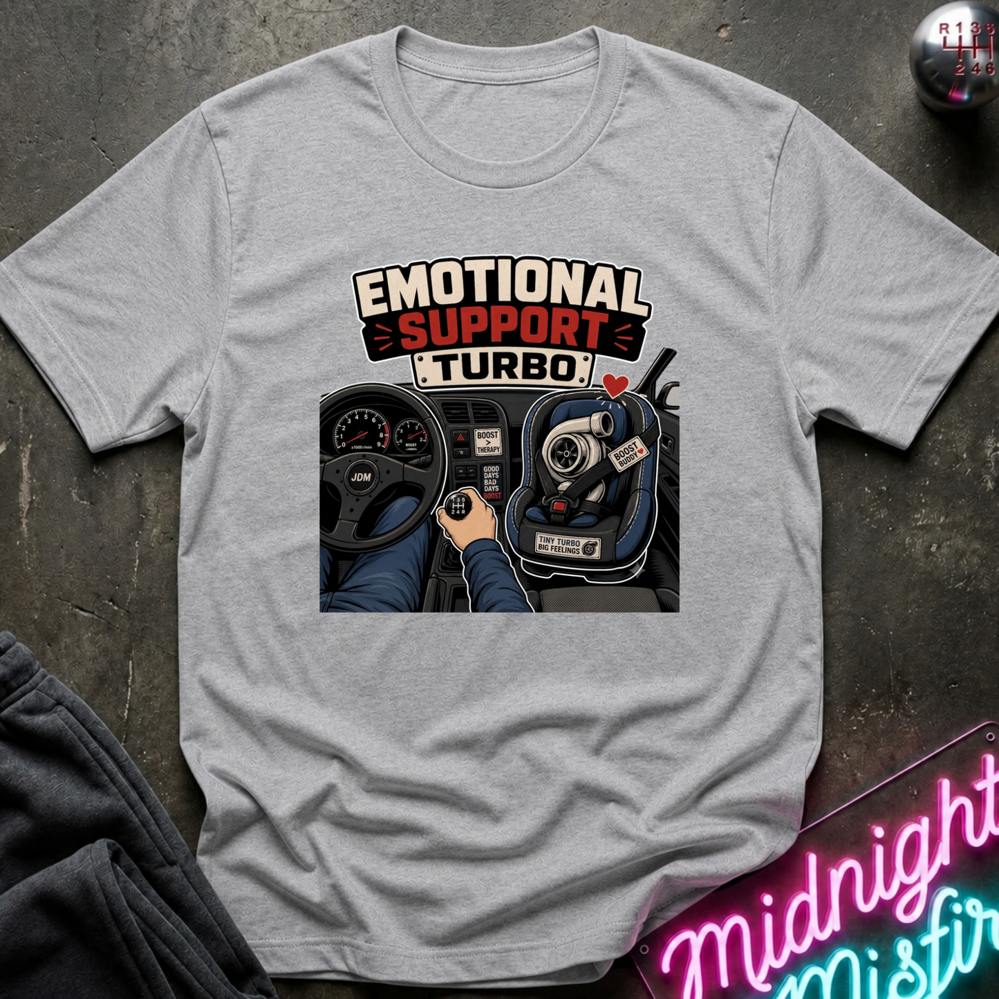 A light gray heather crewneck t-shirt featuring a graphic print of a car dashboard with the text 'EMOTIONAL SUPPORT TURBO' and a hand shifting a manual gear stick, laid flat on a dark textured surface.