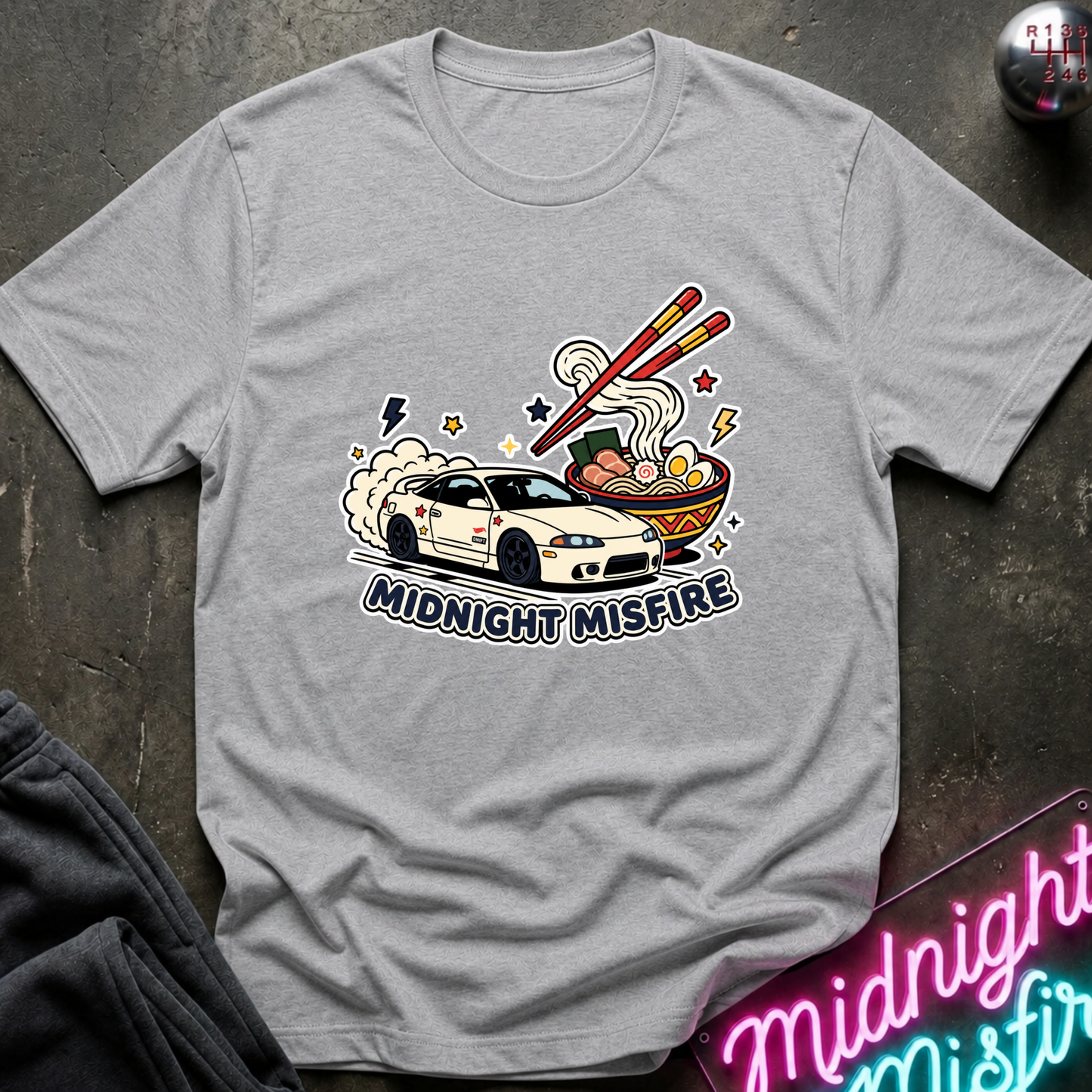 A light gray crewneck t-shirt featuring a graphic of a white sports car with smoke, chopsticks, ramen, and the text 'MIDNIGHT MISFIRE'. The shirt is styled flat on a dark textured surface with a neon sign and dark pants partially visible.