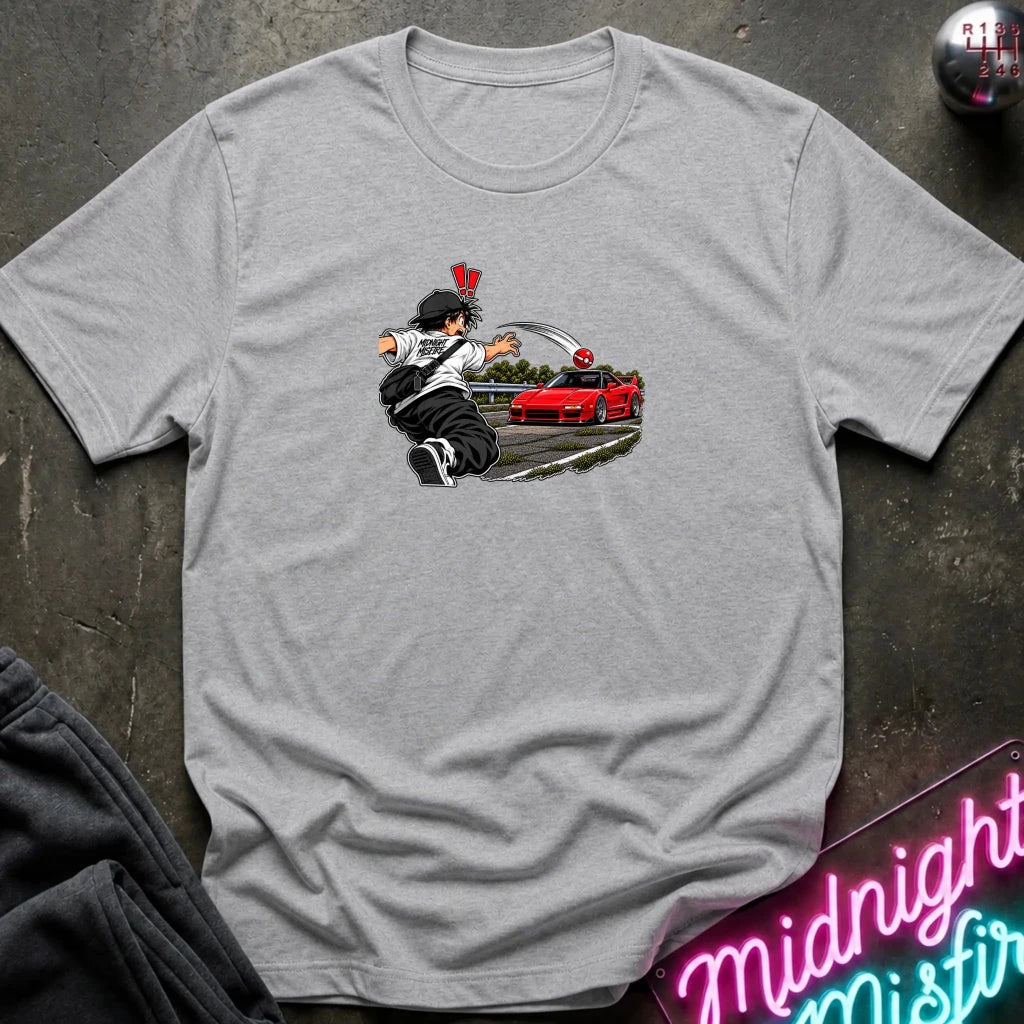 A light gray short-sleeve crewneck t-shirt laid flat on a dark textured surface. The center of the shirt features a graphic print of an anime character in a black and white outfit with a hat, next to a sports car 