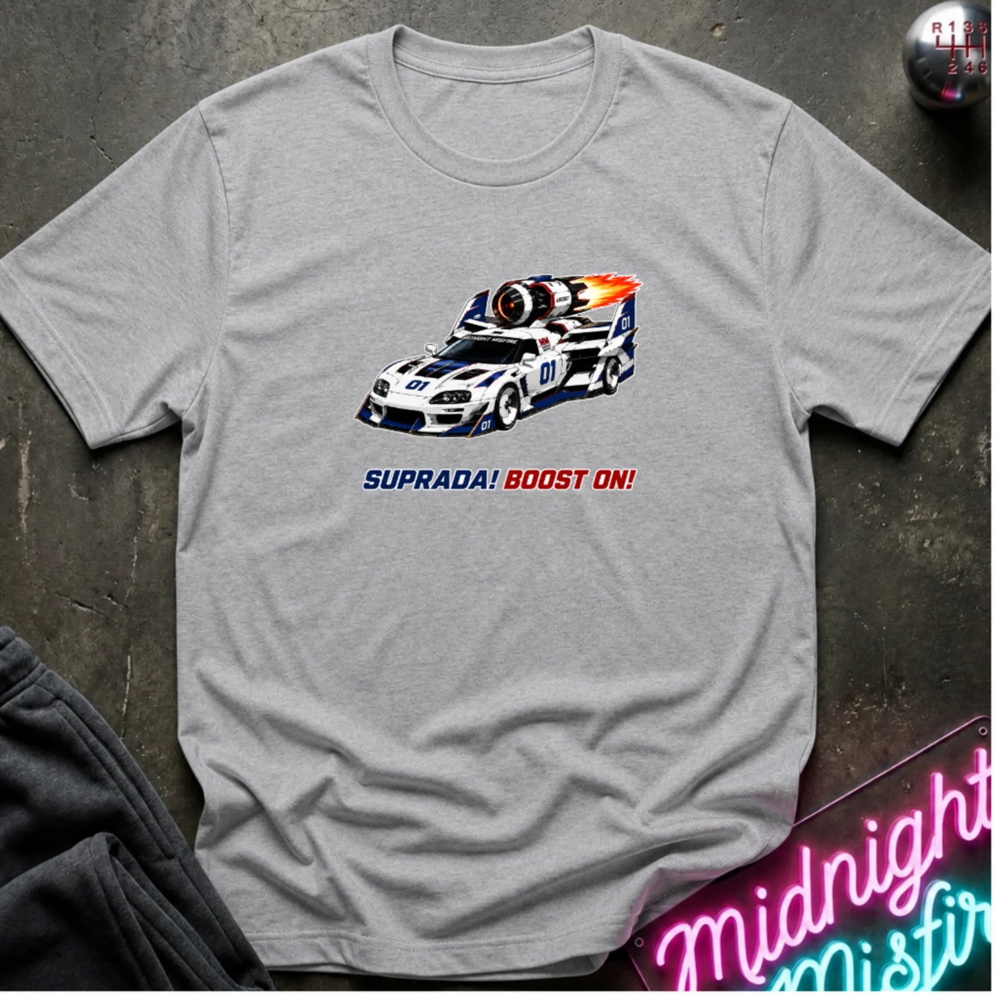 A light gray short-sleeve t-shirt with a retro-style graphic of a race machine with big boost, displayed flat on a dark textured surface with a neon sign and sweatpants partially visible.