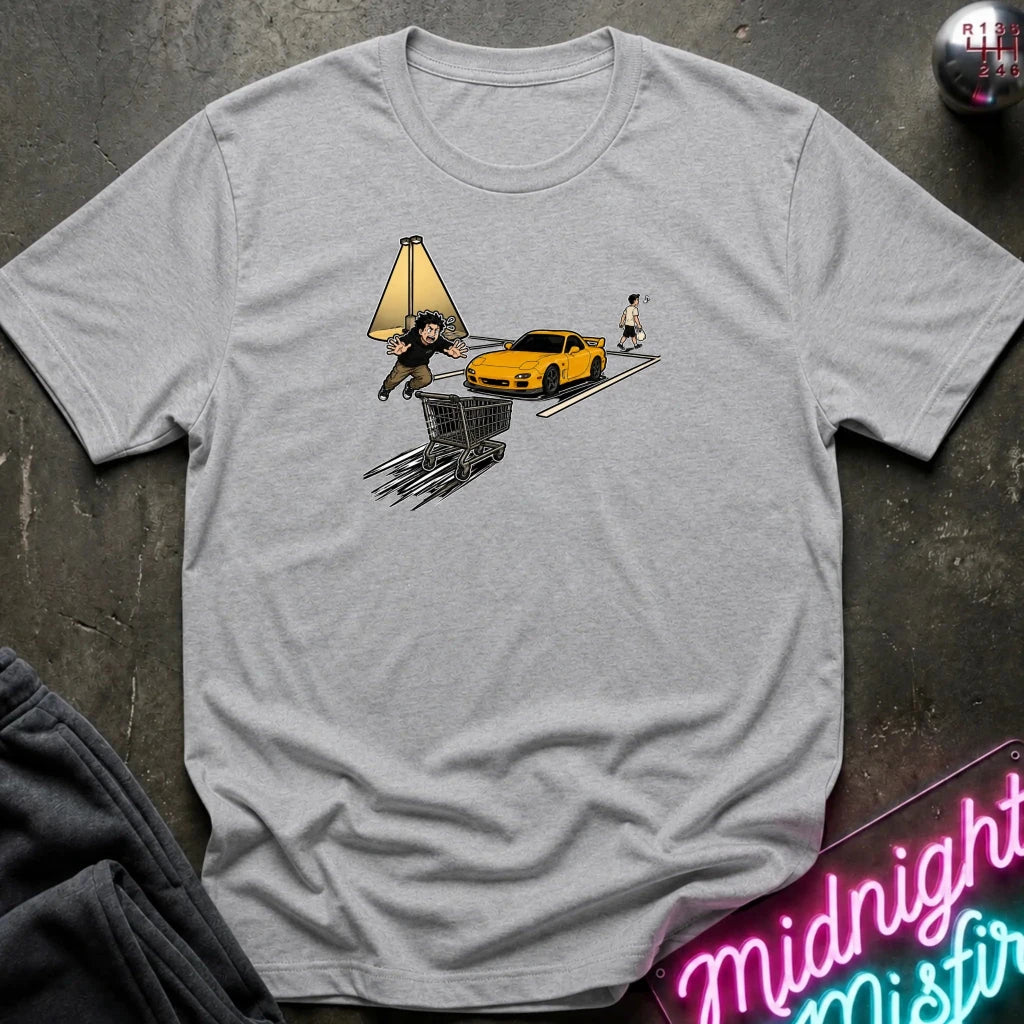 A light heather gray crewneck t-shirt laid flat on a dark textured surface. The shirt features a graphic print of a yellow sports car, a shopping cart, a lamp, and a person, styled with a dark sweatshirt and a neon sign in the corners.