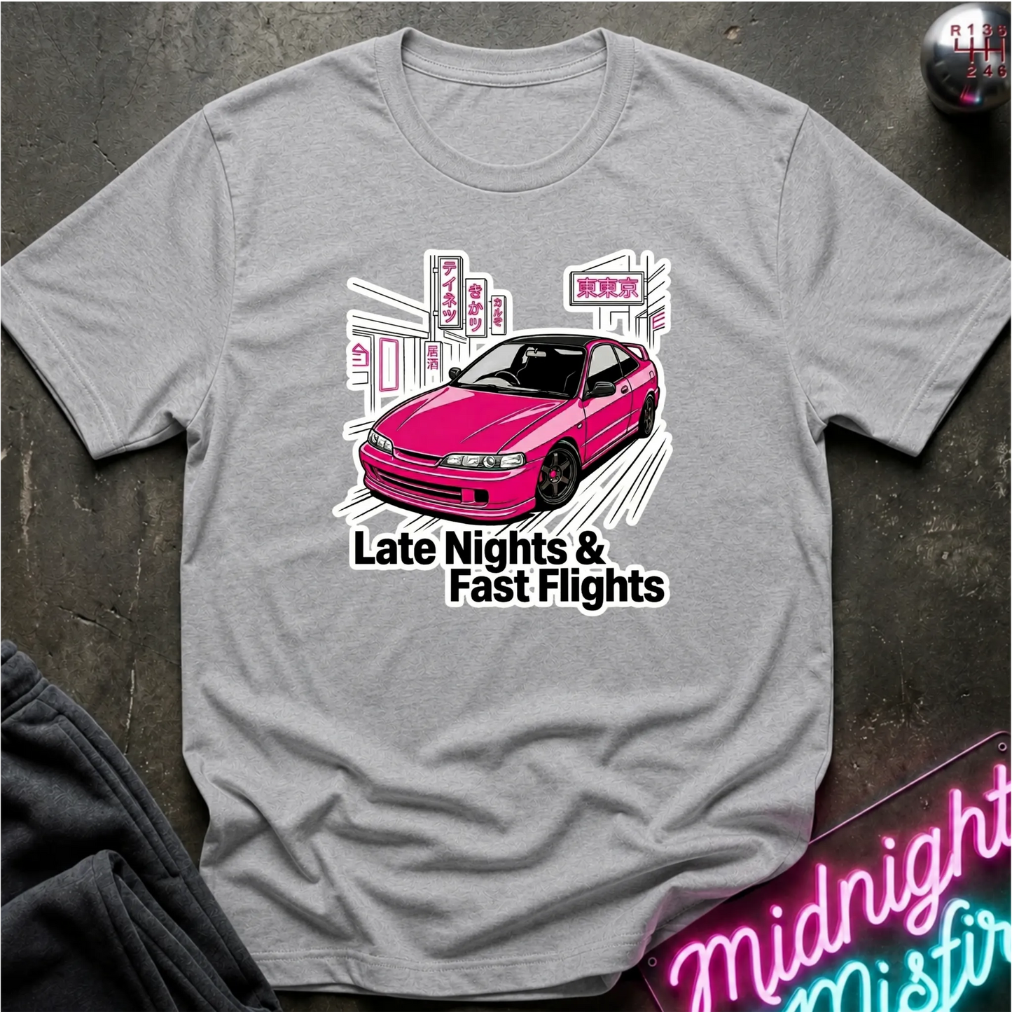A heather gray short-sleeve t-shirt laid flat on a dark textured surface, featuring a graphic of a pink car with the text 'Late Nights & Fast Flights'.
