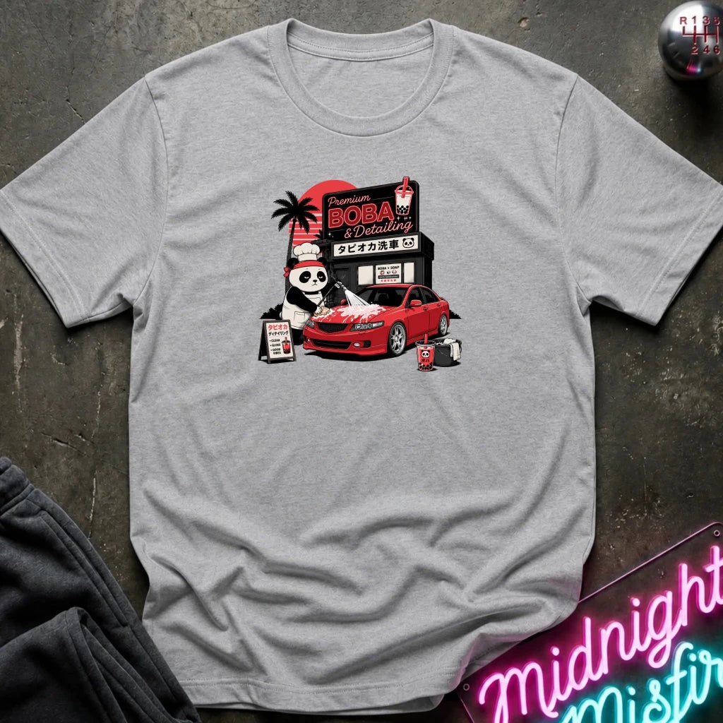 A heather gray short-sleeve graphic t-shirt laid flat on a dark textured surface. The graphic features a cartoon panda wearing a hat next to a red sports car, with a sign reading 'Midnight Boba'.