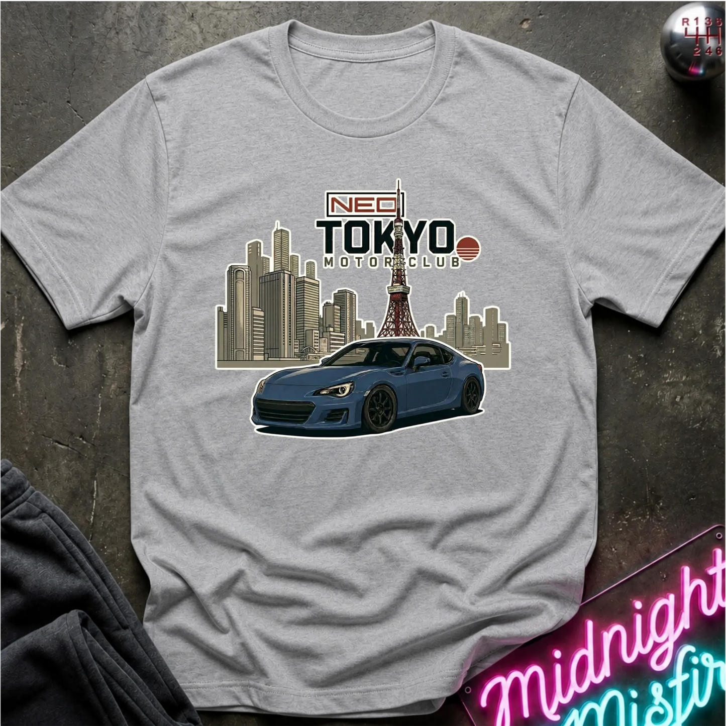 A gray short-sleeve graphic T-shirt laid flat on a dark textured surface. The shirt features a printed design of a blue sports car in front of a Tokyo city skyline with the Tokyo Tower, and text reading 'NEO TOKYO MOTORCLUB'.