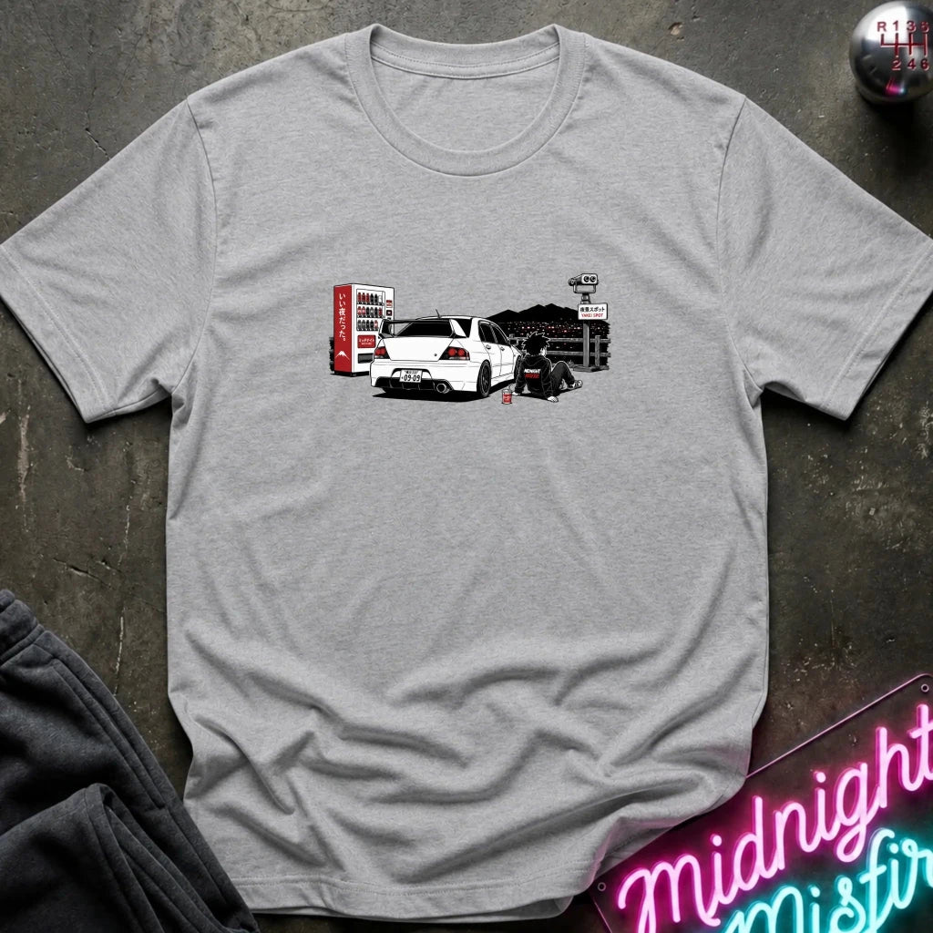 A heather gray short-sleeve t-shirt laid flat on a dark textured surface, featuring a graphic print of a white sports car with a red vending machine.
