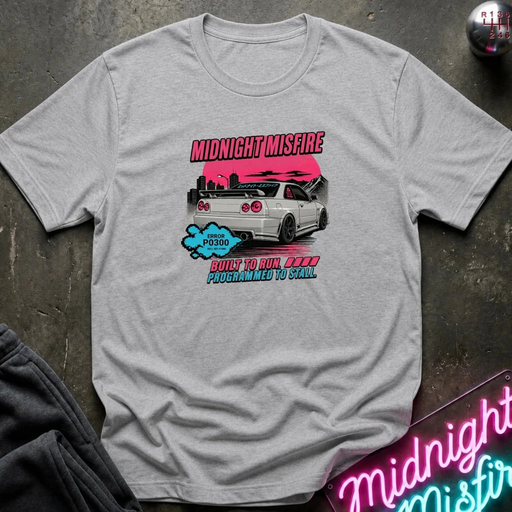 A light gray heather t-shirt laid flat on a dark textured surface, featuring a graphic print of a white sports car with pink, blue, and black accents and the text 'MIDNIGHTMISFIRE'.