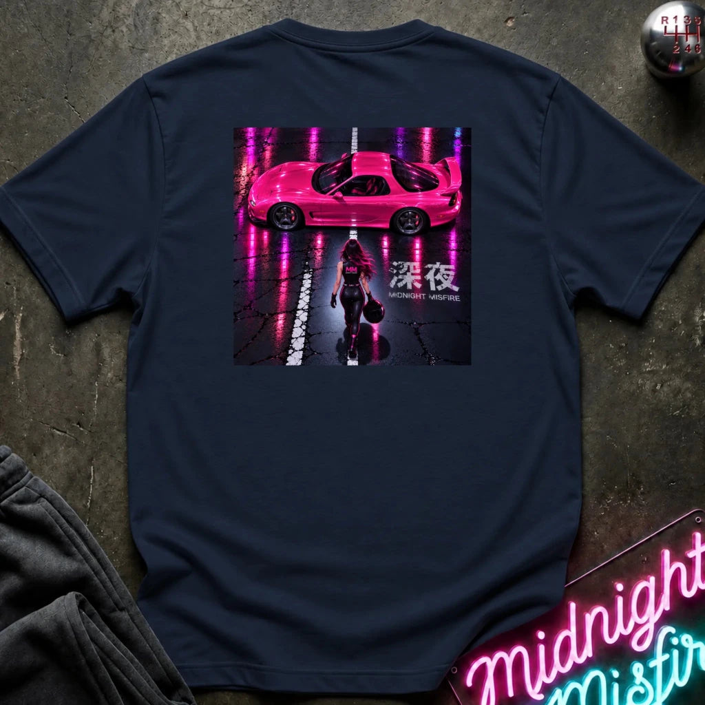 A navy blue t-shirt laid flat on a dark textured surface, featuring a large graphic print on the back of a pink sports car on a wet street at night with a woman walking underneath. The shirt is styled with a gray sweatshirt, a neon sign, and a small metallic ball prop.
