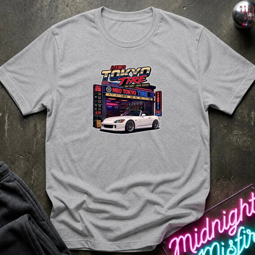 A light gray heather crewneck t-shirt laid flat on a dark textured surface. The shirt features a colorful graphic print in the center depicting a white sports car parked in front of a neon-lit 'Neo Tokyo Tire' garage, with 'NEO TOKYO TIRE' written above it.