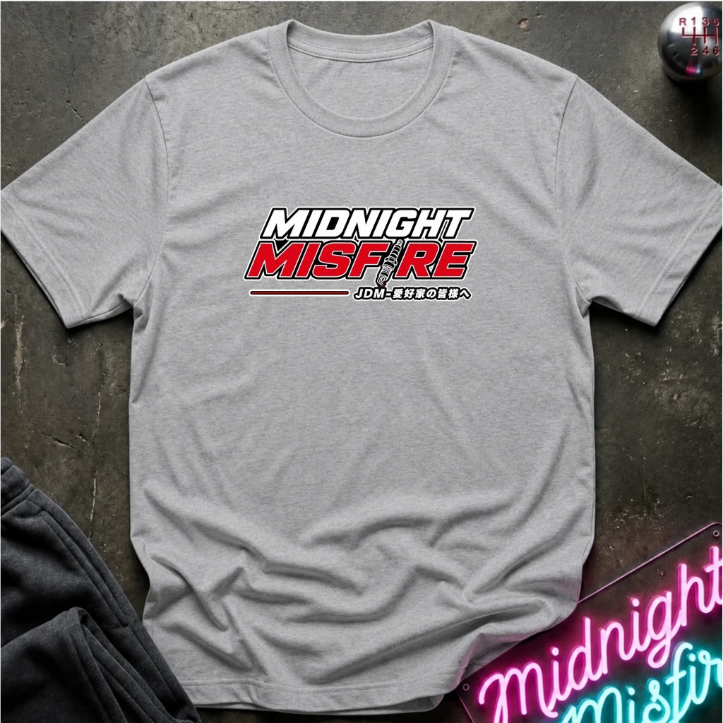 A light gray heather crewneck t-shirt with a 'MIDNIGHT MISFIRE' graphic printed on the chest, laid flat on a dark textured surface with a neon sign and dark pants partially visible.