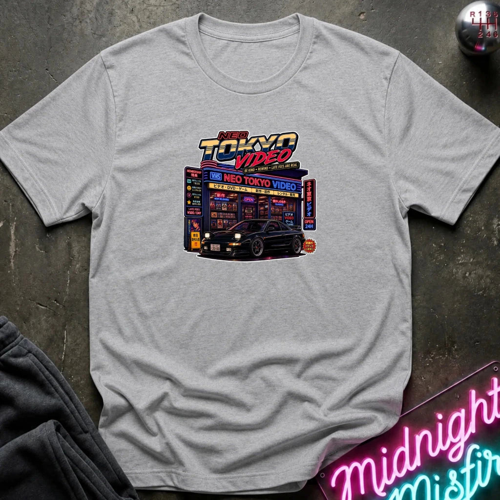 A gray short-sleeve crewneck t-shirt featuring a 'Neo Tokyo video' graphic laid flat on a dark textured surface with a neon sign and sweatpants as props.