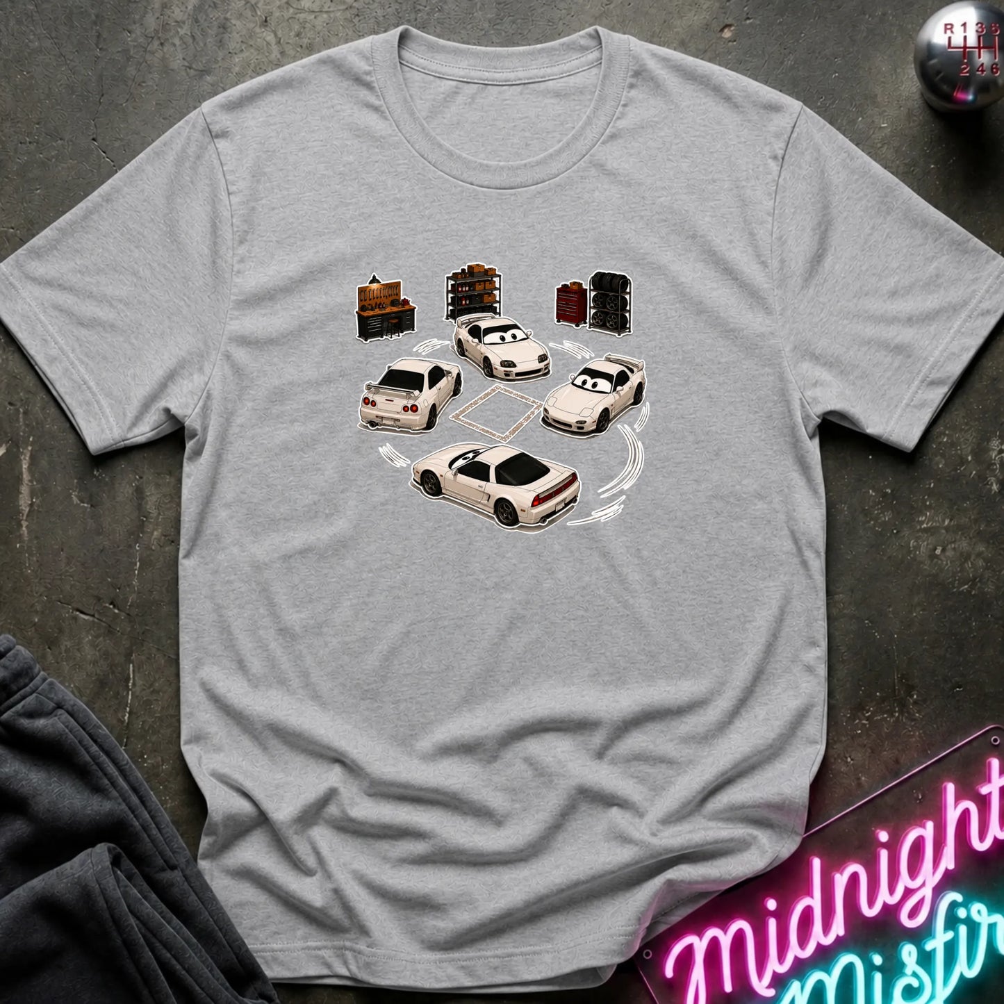 A light gray heather short-sleeve t-shirt laid flat on a dark textured surface, featuring a graphic print of several white sports cars arranged in a circle with drift-like tire marks.