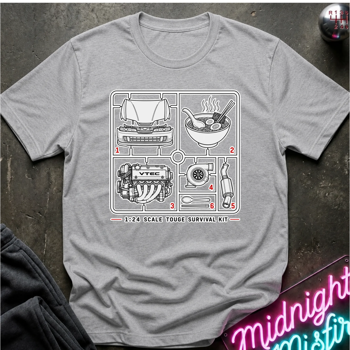 A heather gray short-sleeve graphic T-shirt laid flat on a dark textured surface. The shirt features a black line-art schematic of a survival kit with numbered illustrations and the text '1:24 SCALE TOUGHE SURVIVAL KIT' printed on the chest.
