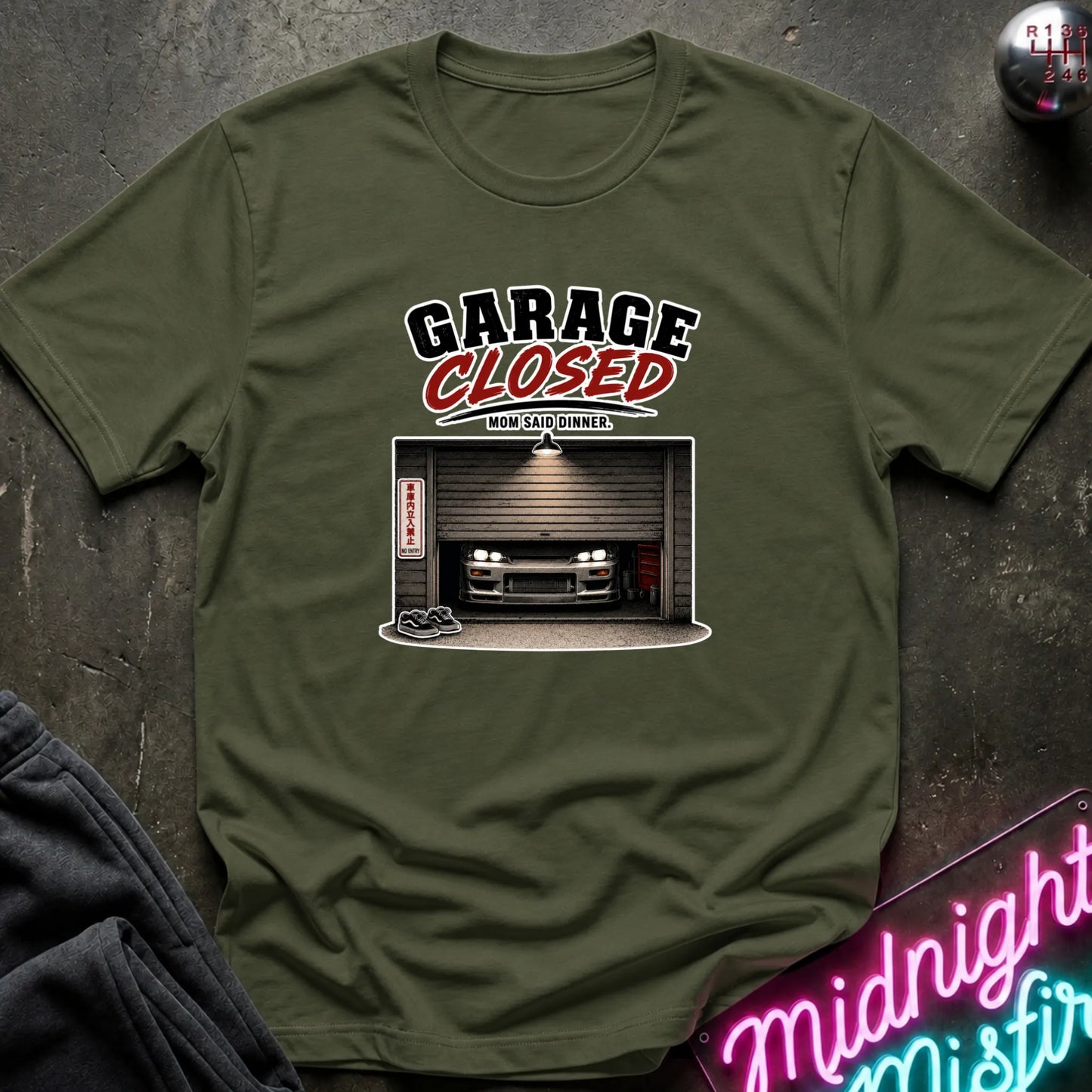 An olive green short-sleeve t-shirt laid flat on a dark textured surface, featuring a graphic print that reads 'GARAGE CLOSED' above an illustration of a car parked inside a closed garage. A neon sign and folded dark fabric are partially visible around the shirt.