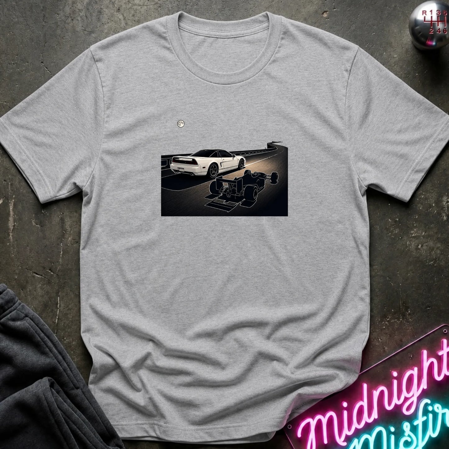 A light gray heather short-sleeve crewneck t-shirt laid flat on a dark textured surface, featuring a graphic print of a white sports car and a shadow car on a night road scene. The shirt is styled with dark gray sweatpants, a silver ball, and a neon sign in the corners.