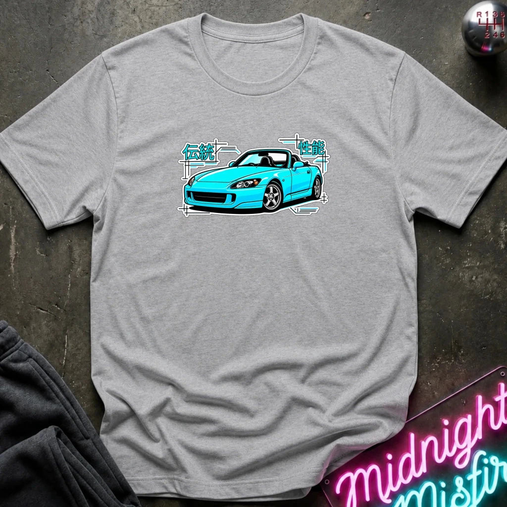 A light heather gray t-shirt laid flat on a dark textured surface, featuring a graphic print of a bright blue sports car with white and teal text elements. A neon sign and dark clothing are partially visible in the styled flat-lay composition.