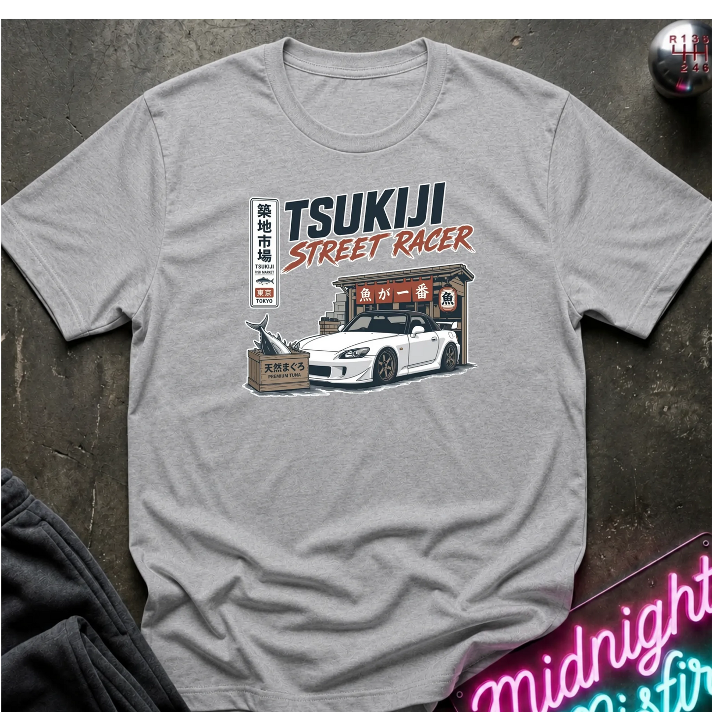 Gray short-sleeve graphic T-shirt laid flat on and surrounding an illustration of a white sports car parked outside a fish market.