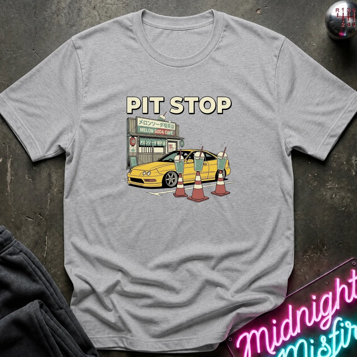 A heather gray short-sleeve t-shirt with a cartoon graphic of a yellow car at a 'Pit Stop' cafe, surrounded by traffic cones. The shirt is laid flat on a dark textured surface with a neon sign and dark clothing partially visible.