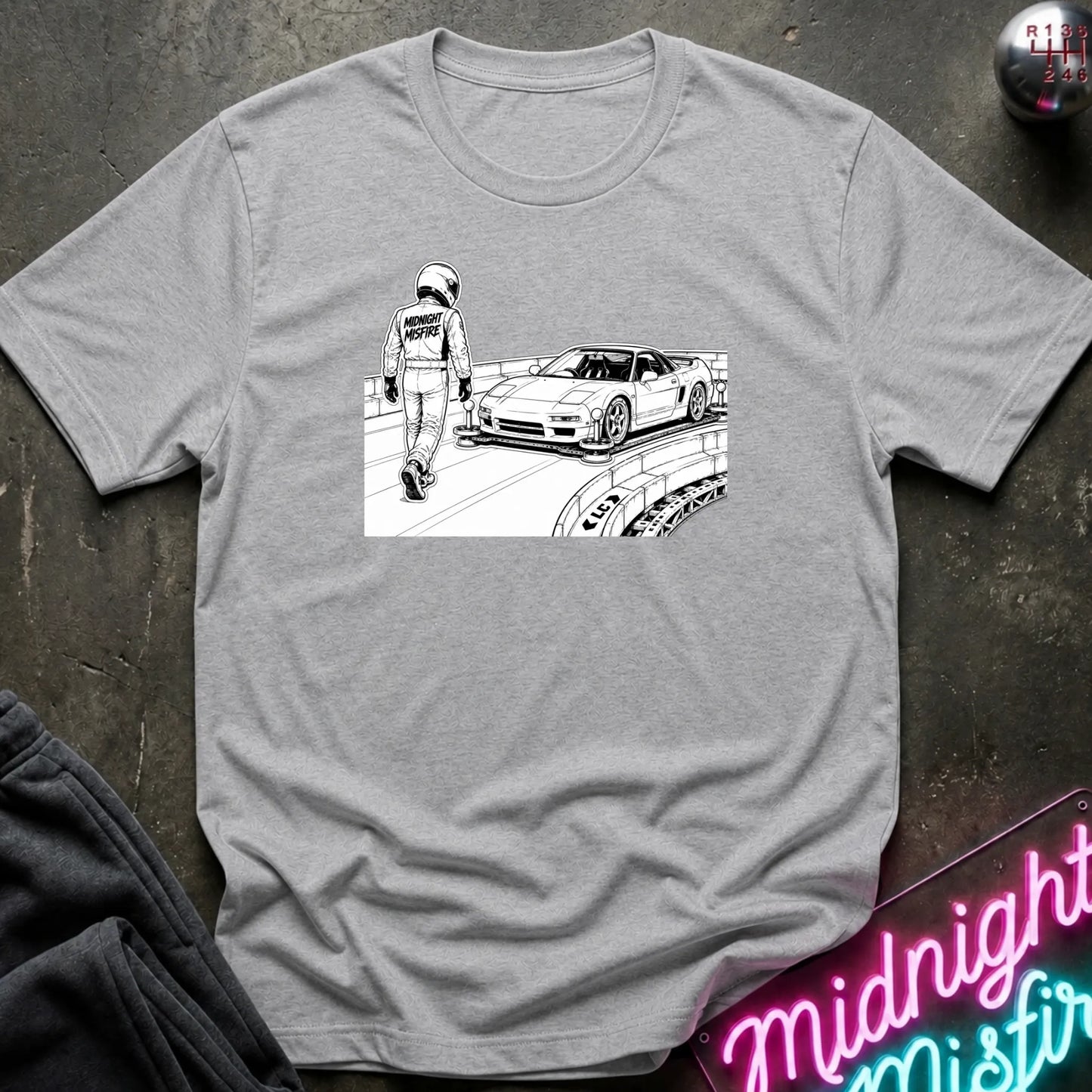 A heather gray short-sleeve t-shirt with a black and white graphic print of a race car and a driver on the chest, displayed flat on a dark textured surface with lifestyle props.