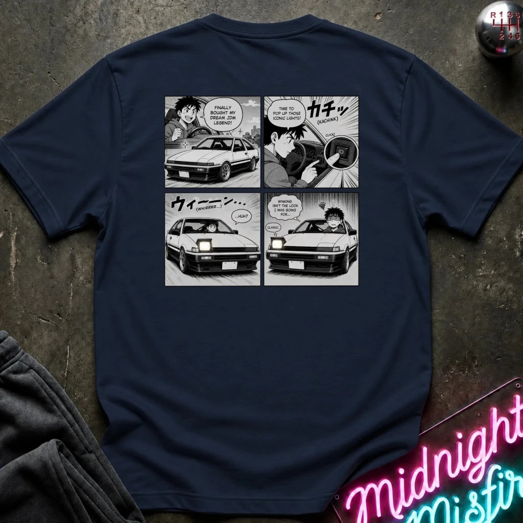 A navy blue t-shirt laid flat on a dark textured surface, featuring a black and white manga comic strip graphic on the back showing characters in a car. 
