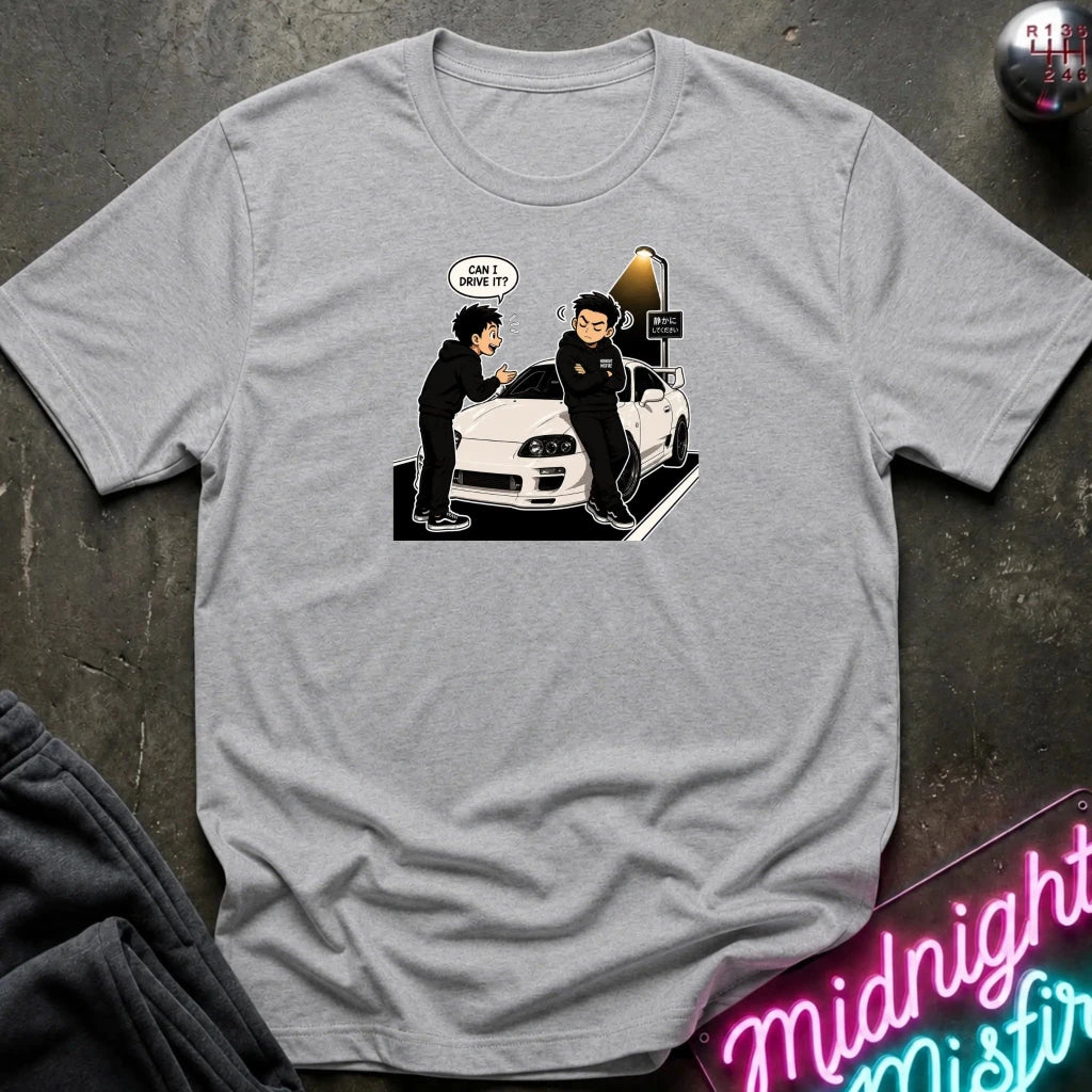 A heather gray short-sleeve t-shirt laid flat on a dark textured surface, featuring a cartoon graphic of two anime-style characters standing next to a white sports car with a speech bubble that reads 'CAN I DRIVE IT?'. 