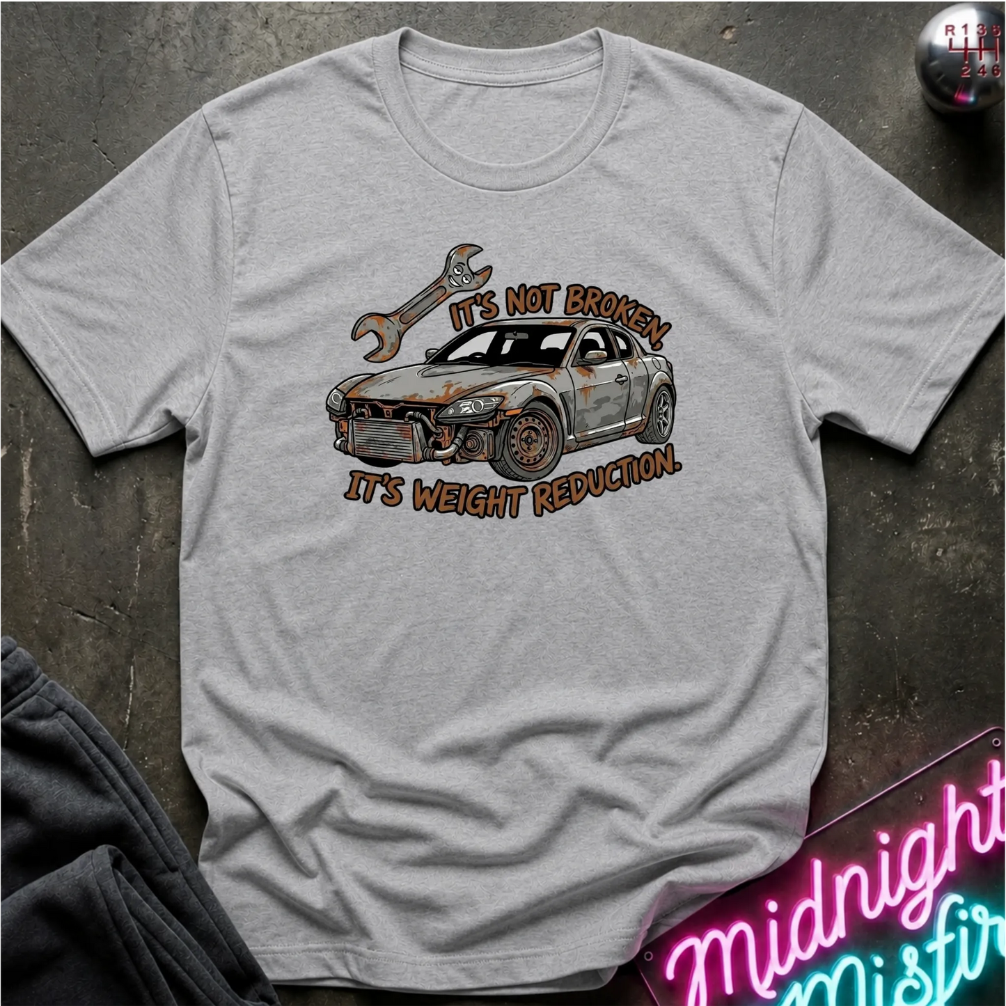 A light gray crewneck t-shirt featuring a graphic of a rusted-out car with a wrench and the text 'IT'S NOT BROKEN IT'S WEIGHT REDUCTION'. The shirt is laid flat on a dark textured surface with a neon sign and dark pants partially visible.