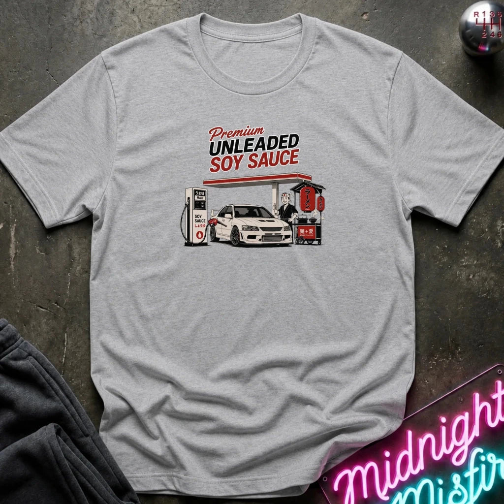 A light gray heather crewneck t-shirt laid flat on a dark textured surface. The shirt features a graphic print of a white sports car at a gas station with the text 'Premium UNLEADED SOY SAUCE' in red and black.