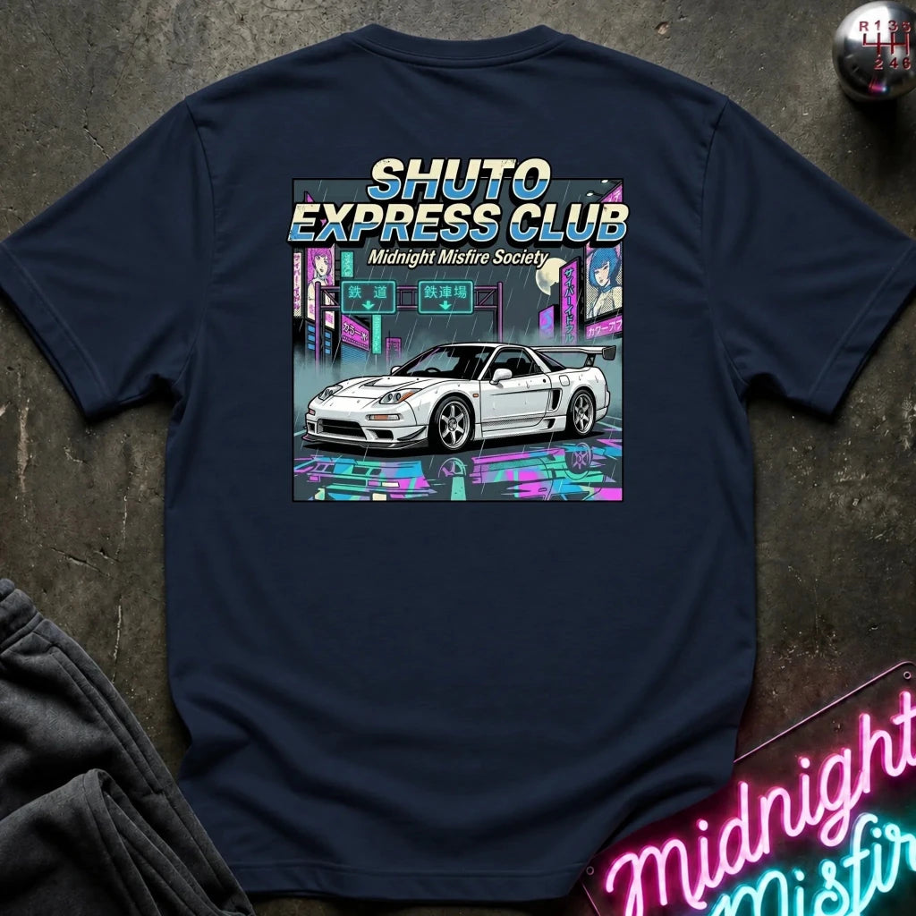 A navy blue t-shirt laid flat on a dark textured surface, featuring a large graphic print on the back with the text 'SHUTO EXPRESS CLUB' and 'Midnight Misfire Society' above an illustration of a white sports car in a neon city scene. 