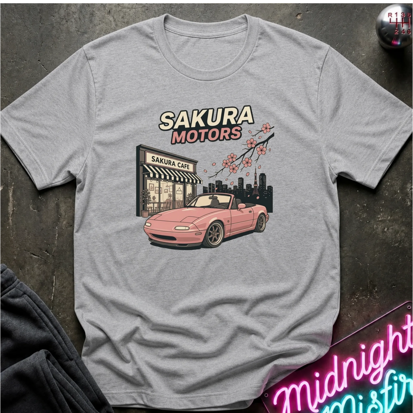 A light gray crewneck t-shirt laid flat on a dark textured surface, featuring a graphic print of a pink convertible car in front of a 'Sakura Motors' cafe with cherry blossoms.
