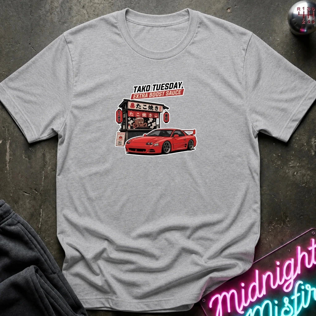 A light gray crewneck t-shirt laid flat on a dark textured surface, featuring a graphic print of a red sports car and a Japanese street food stall with the text 'TAKO TUESDAY'.