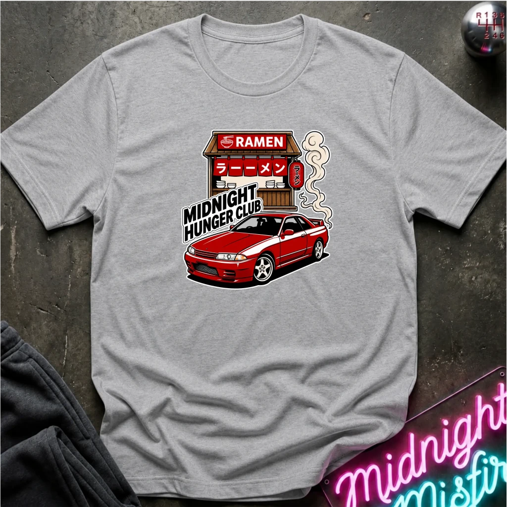 A light gray crewneck t-shirt featuring a graphic print of a red sports car and a ramen shop with the text 'MIDNIGHT HUNGER CLUB'. The shirt is displayed flat on a dark textured surface with a neon sign and dark fabric partially visible in the corners.