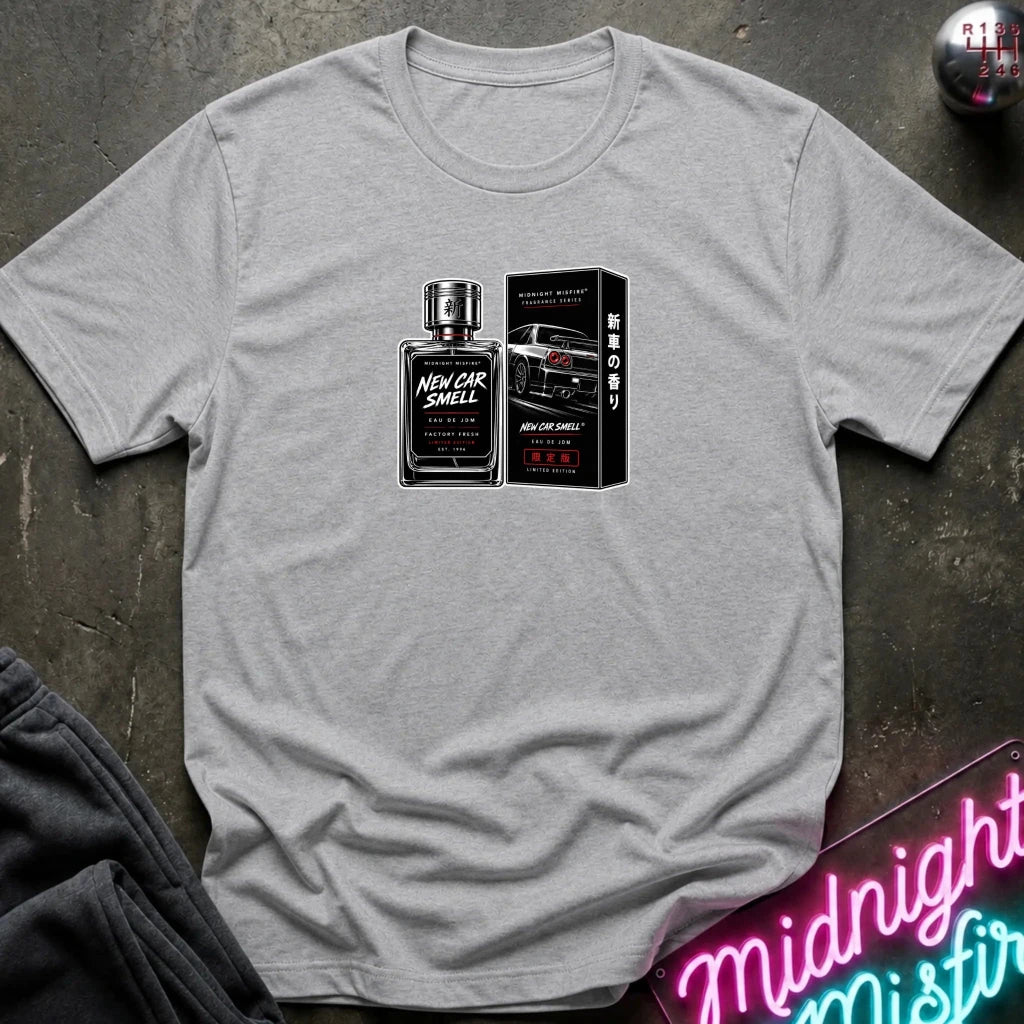 A light gray heather crewneck t-shirt laid flat on a dark textured surface, featuring a graphic print of a perfume bottle and box labeled 'New Car Smell' with a sports car. 