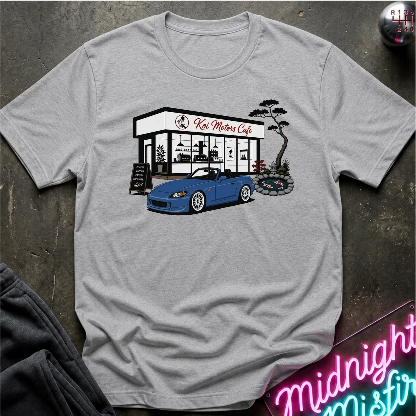 A light gray heather crewneck t-shirt featuring a graphic print of a blue convertible sports car parked outside 'Koi Motor Cafe'. The shirt is displayed flat on a dark textured surface with a neon sign and dark pants partially visible in the corners.